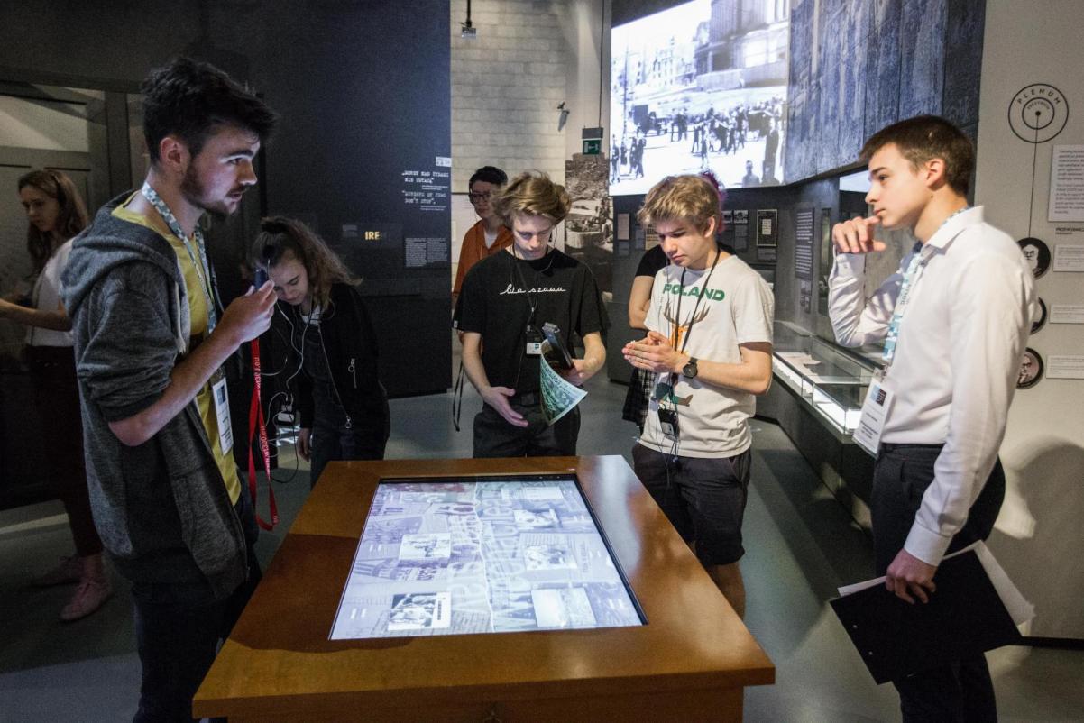 Grupa młodzieży ze słuchawkami na uszach, stoi wokół gabloty, w której wyświetlane są zdjęcia. gablota znajduje się w przestrzeni wystawy stałej Muzeum POLIN