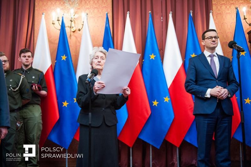 Mateusz Morawiecki podczas konferencji. W tle polskie i unijne flagi. W lewym dolnym rogu logo POLIN i portalu Polscy Sprawiedliwi.
