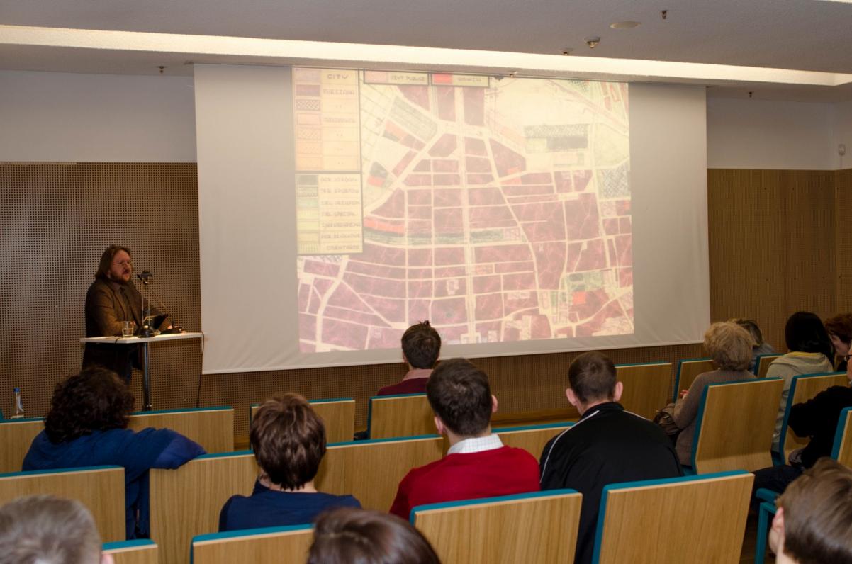Paweł Weszpiński stoi przy pulpicie podczas wykładu, na ekranie wyświetla się slajd prezentacji.