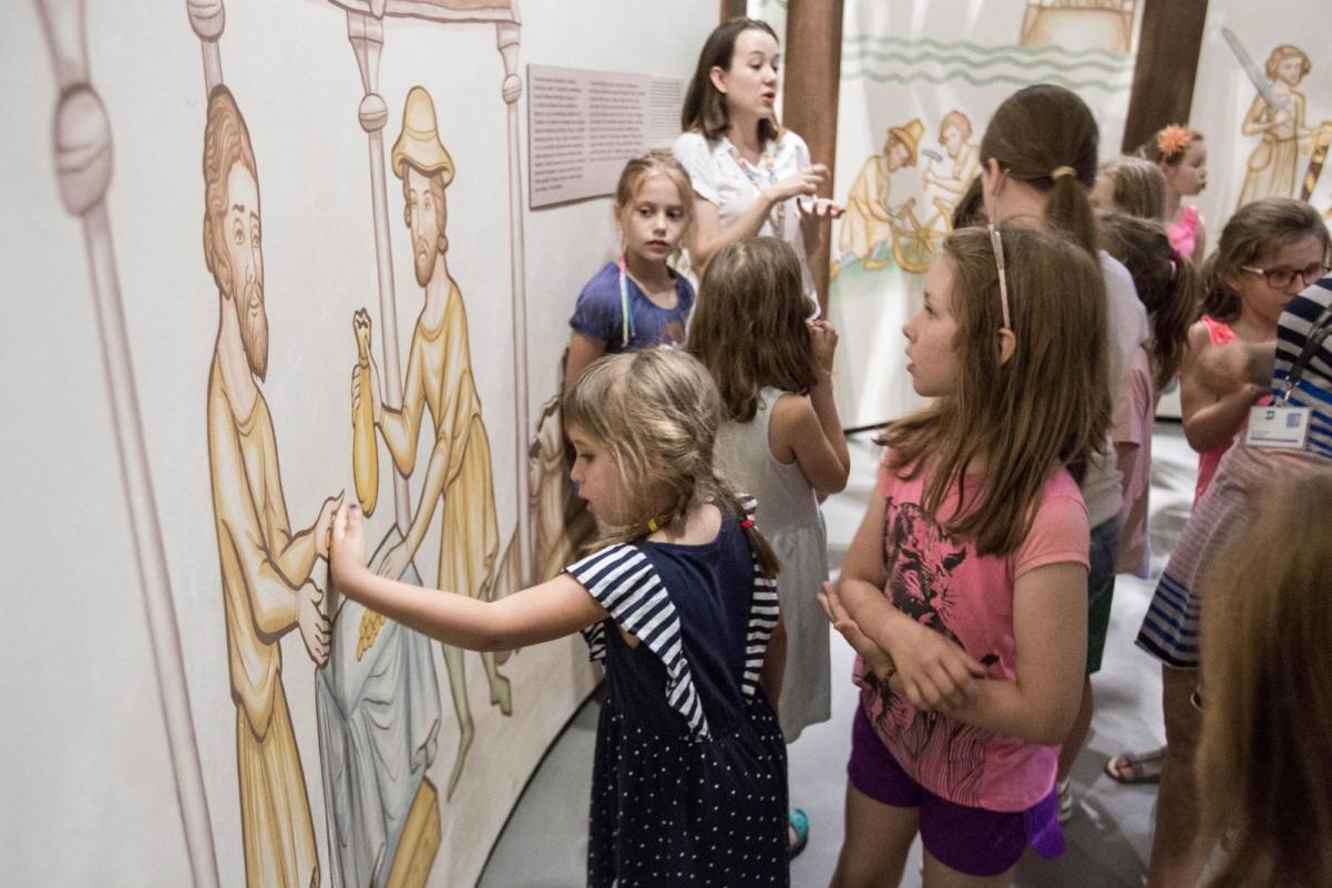 Warsztaty dla klas 1-3 w Muzeum POLIN. Grupa dzieci stoi przed rysunkiem na ścianie, przedstawiającym dawnego rzemieślnika. Jedno z dzieci dotyka rysunku. Z tyłu, za dziećmi stoi kobieta z ciemnymi włosami do ramion.
