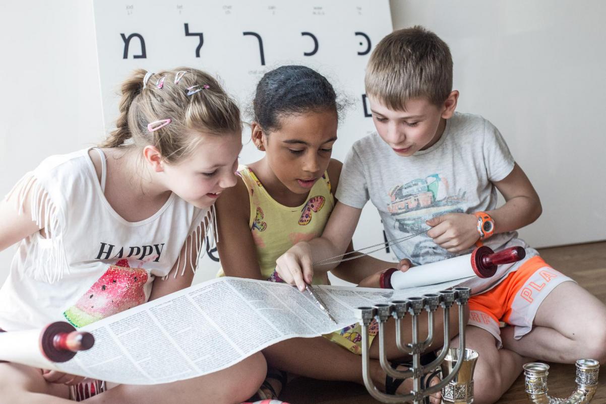 Troje dzieci pochyla się nad dużym arkuszem papieru, za nimi na ścianie litery alfabetu hebrajskiego - Warsztaty dla szkół podstawowych klas 1-3 w Muzeum POLIN