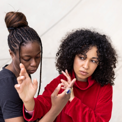 Dwie ciemnoskóre kobiety stoją obok siebie. Trzymają dłonie w górze, w pobliżu swoich twarzy.