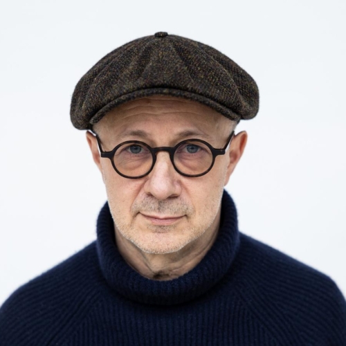 Mikołaj Grynberg - mężczyzna ubrany na czarno, na nosie ma okulary w ciemnych oprawkach, a na głowie szary kaszkiet.