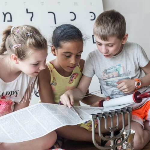 Troje dzieci ogląda zwój Tory na warsztatach edukacyjnych. Za nimi stoi tablica z alfabetem hebrajskim.