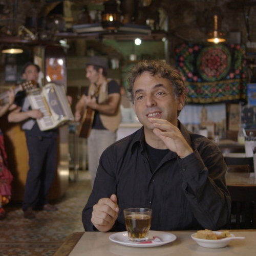 Kadr z filmu "Etgar Keret. Oparte na prawdziwej historii". Keret siedzi przy stole, pije herbatę, w tle zespół z instrumentami.