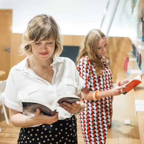 Dwie kobiety stoją przy regale z książkami w bibliotece. Obie czytają jakieś publikacje. / Two women are standing by a bookcase in a library. They are both reading some publications.