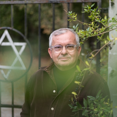 Andrzej Białek widziany zza zielonych krzewów. Za nim ogrodzenie żydowskiego cmentarza z gwiazdą Dawida