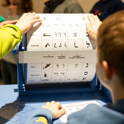 Chłopiec obraca wałek edukacyjny z nazwami słów i literami w różnych językach żydowskich.