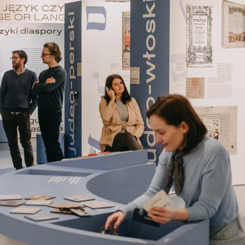 На першым плане дзяўчына раскладвае карткі на выставе. Другая - слухае аўдыягід. На другім плане стаяць два мужчыны, якія знаёмяцца з экспазіцыяй.