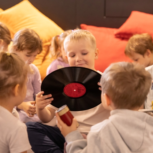 Chłopiec trzyma płytę winylową. Dookoła niego siedzą inne dzieci. / The boy is holding a vinyl record. Other children are sitting around him.