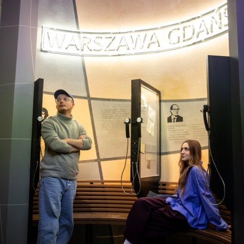 Mężczyzna stoi z założonymi rękami, a kobieta siedzi na ławce na wystawie stałej w Muzeum POLIN.