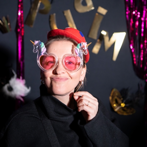 Kobieta w różowych okularach z jednorożcami i czerwonej czapce. W tle ścianka ze złotym napisem Purim i kolorowymi serpentynami.