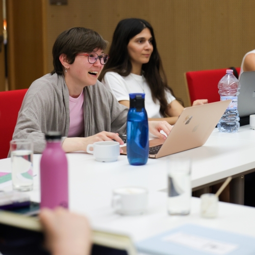 A group of women are sitting at a table in a conference room. They are holding laptops with open screens in front of them.