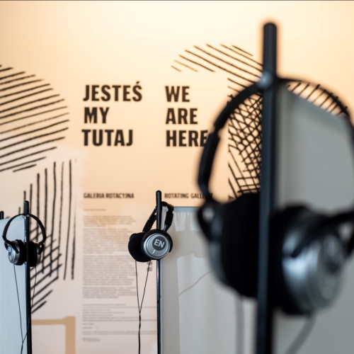 Słuchawki wiszą na kołkach w Galerii Rotacyjnej. Na ścianie napisy: jesteśmy tutaj, we are here.