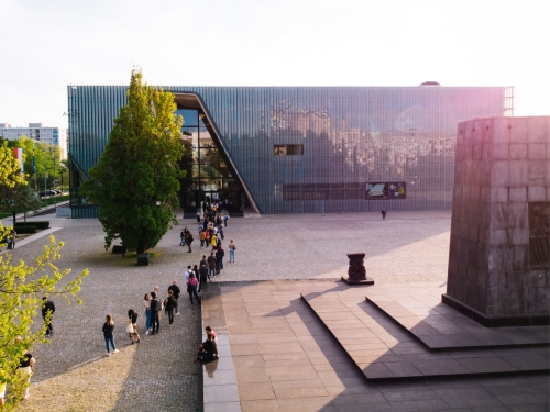 Kolejka osób czeka na wejście do budynku Muzeum POLIN. Świeci słońce. Z prawej strony widoczny pomnik Bohaterów Getta. / A queue of people is waiting to enter the POLIN Museum building. The sun is shining. The Monument to the Ghetto Heroes is visible on the right.