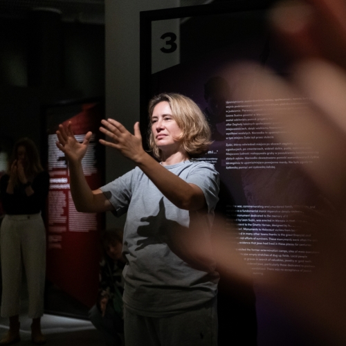 In the foreground, blurred hands. In the background, a woman with her hands raised.