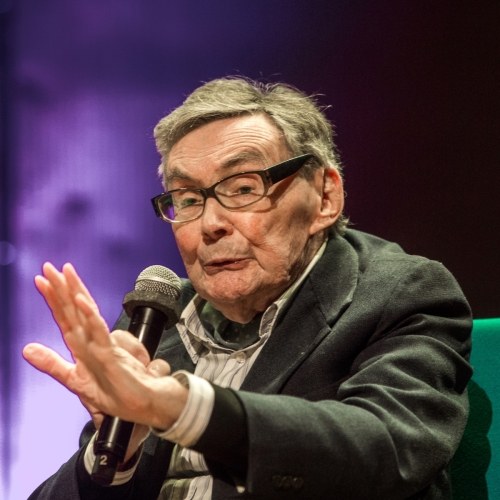 Marian Turski mówi przez mikrofon i wznosi jedną dłoń ku górze.