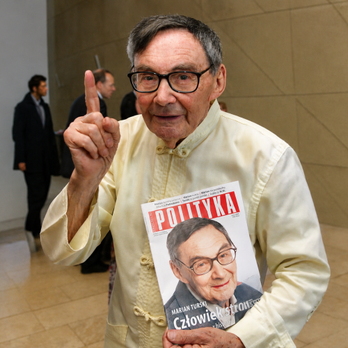 Marian Turski trzyma przed sobą egzemplarz Polityki i wznosi palec ku górze.