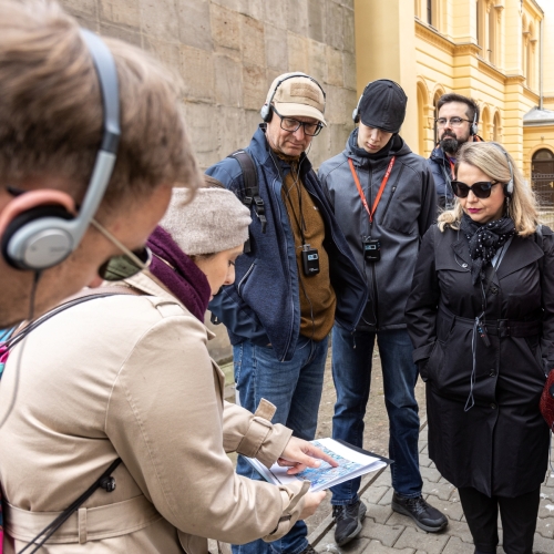 Grupa uczestniczek i uczestników spaceru miejskiego po Warszawie. Przewodniczka pokazuje im coś palcem w swoich notatkach.