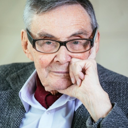 Marian Turski ubrany w niebieską koszulę i marynarkę. Podpiera twarz dłonią i patrzy przed siebie.