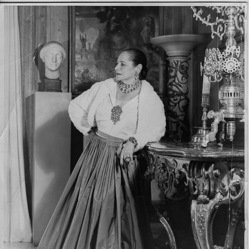 Helena Rubinstein w białej bluzce z bufiastymi rękawami i długiej spódnicy, stoi oparta o ozdobną komodę. W tle dzieła sztuki.